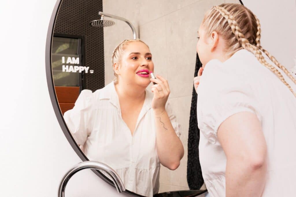 A woman applies pink lipstick in front of a mirror as part of Liptember.