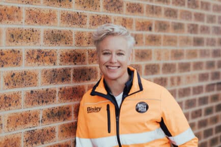 Jo Farrell standing against brick wall with high-vis orange jacket